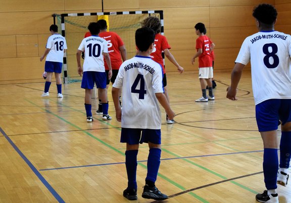 Das Bild zeigt fußballspielende Kinder in einer Turnhalle, sie tragen blau-weiße Trikots.