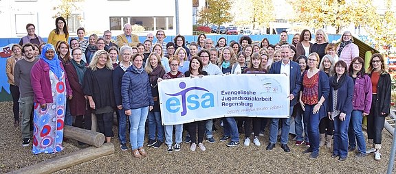 Das Bild zeigt eine Gruppe von Menschen auf einem Spielplatz, die ein Banner der ejsa Regensburg halten.