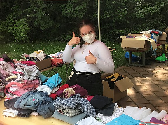 Das Bild zeigt eine junge Frau mit Maske, die hinter einem Tisch mit Klamotten steht und die Daumen nach oben streckt.