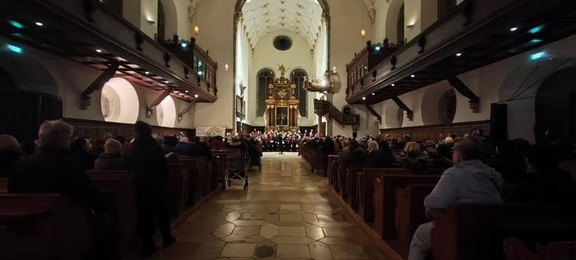 Quelle: Sabine Wunderlich Das Bild zeigt den Innenraum einer Kirche, vor dem Altar steht eine Chorformation, die Reihen sind gefüllt mit Publikum.