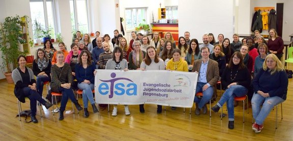 Das Bild zeigt eine Gruppe von Menschen, die in einer Gruppe beisammen sitzen und ein Banner hochhalten.