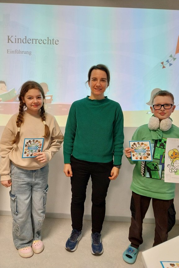 Das Bild zeigt zwei Kinder, die ein ausgemaltes Bild zeigen. In deren Mitte steht eine Frau.  