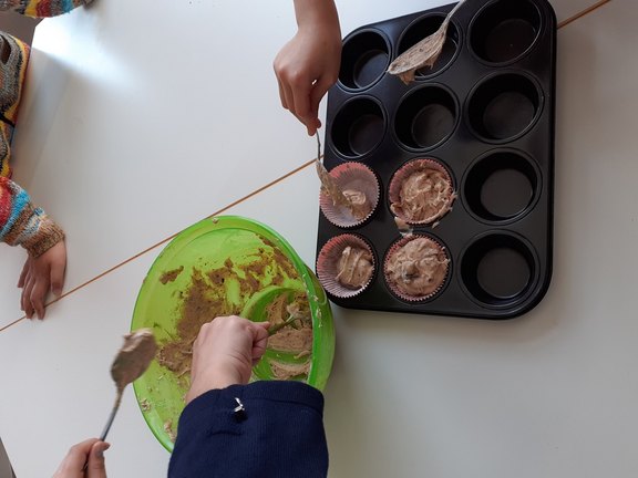 Das Bild zeigt eine Muffinform, in die mehrere Hände gerade Teig aus einer Schüssel füllen.