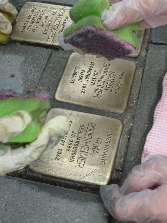 Das Bild zeigt Stolpersteine, am Rand sind Hände zu sehen, die einen Putzschwamm halten.