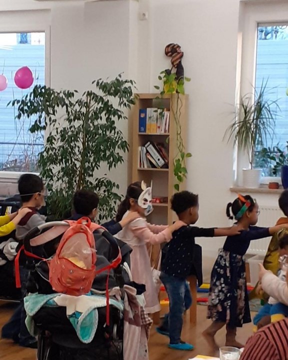 Das Bild zeigt Kinder, die ihre Arme auf die Schultern des jeweiligen Kindes in der Reihe zuvor legen und so als Polonaise durch die Räume ziehen.