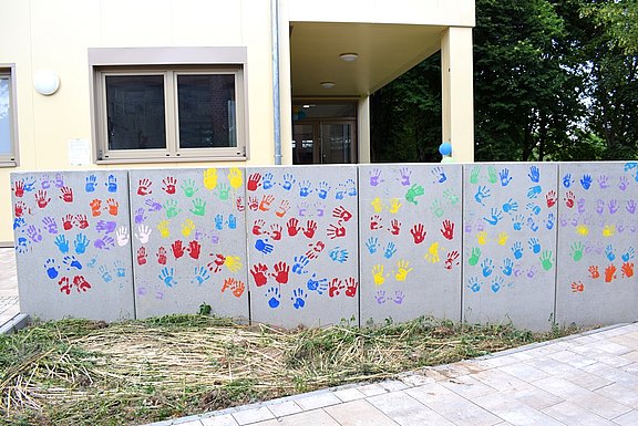 Das Bild zeigt eine graue Mauer mit bunten Handabdrücken darauf.  