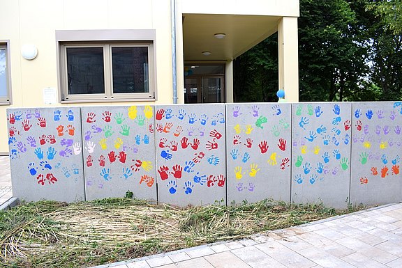 Das Bild zeigt eine graue Mauer mit bunten Handabdrücken darauf.