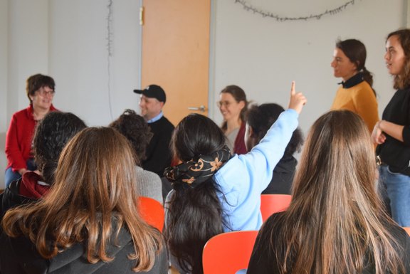 Das Bild zeigt ein Kind, das sich meldet von hinten. Im Hintergrund sind mehrere Personen zu sehen.  