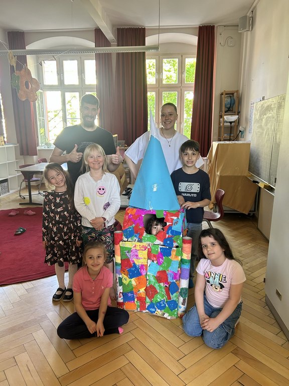 Das Bild zeigt eine Gruppe aus Kindern und Erwachsenen, die um eine Rakete aus Pappkartons versammelt stehen.
