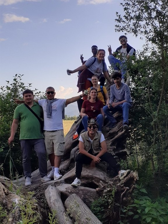 Das Bild zeigt eine Gruppe von 9 Personen, die für ein Foto posieren. Sie haben sich auf einer großen Baumwurzel im Wald platziert und lächeln in die Kamera.