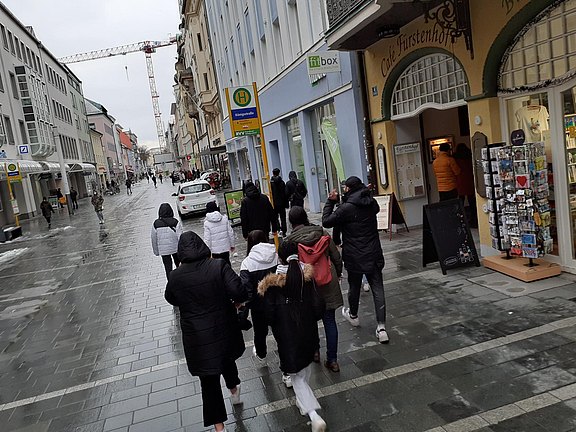 Das Bild zeigt eine Gruppe Jugendlicher von hinten, die durch eine Straße gehen, vorbei an Geschäften usw.