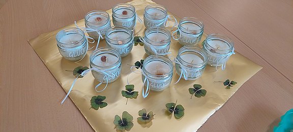 Das Bild zeigt mehrere Kerzen in einem Glas, auf denen das Wort Glück zu lesen ist, sowie vierblättrige Kleeblätter.  