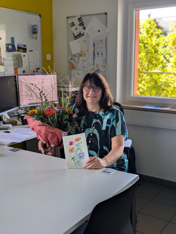 Das Bild zeigt eine Frau, die an einem Schreibtisch sitzt und einen Blumenstrauß in der Hand hält.