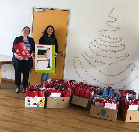 Zwei Frauen stehen vor einer Wand mit einem Baum aus Lichterketten. Vor Ihnen liegen Geschenke.