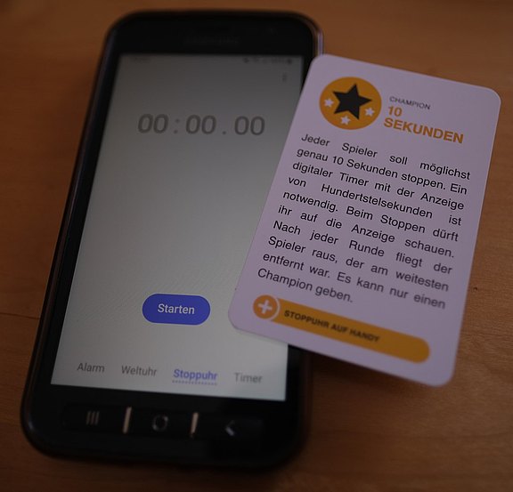 Das Bild zeigt eine Karte mit einer Aufgabe und ein Smartphone mit Stoppuhr.