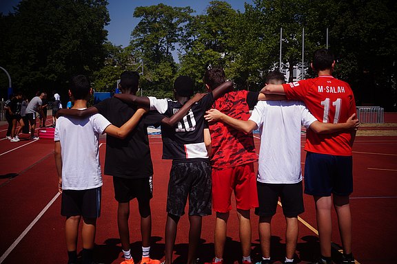 Das Bild zeigt Jugendliche in Sporttrikots, Arm in Arm von hinten.  