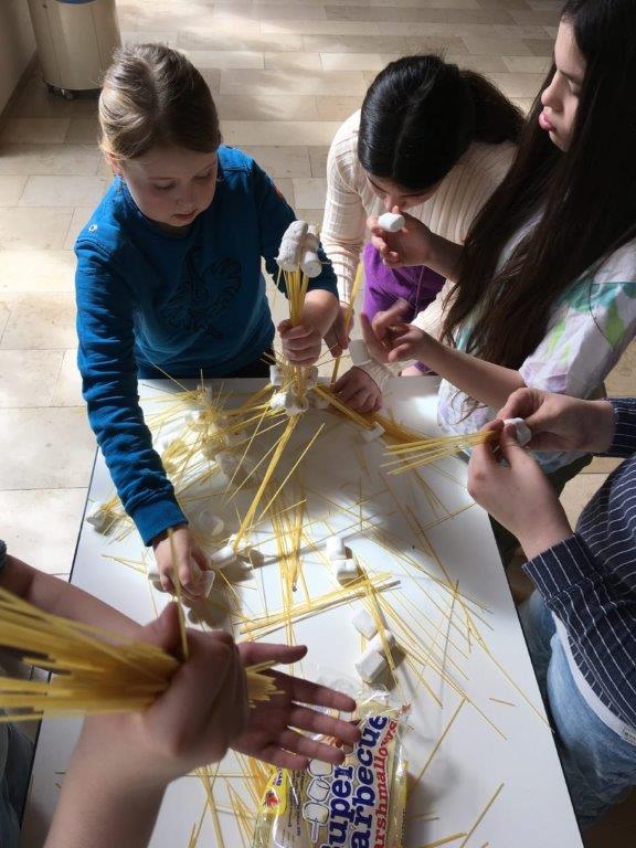 Das Bild zeigt mehrere Kinder, die um einen Tisch herumstehen und mit Spaghettis experimentieren.